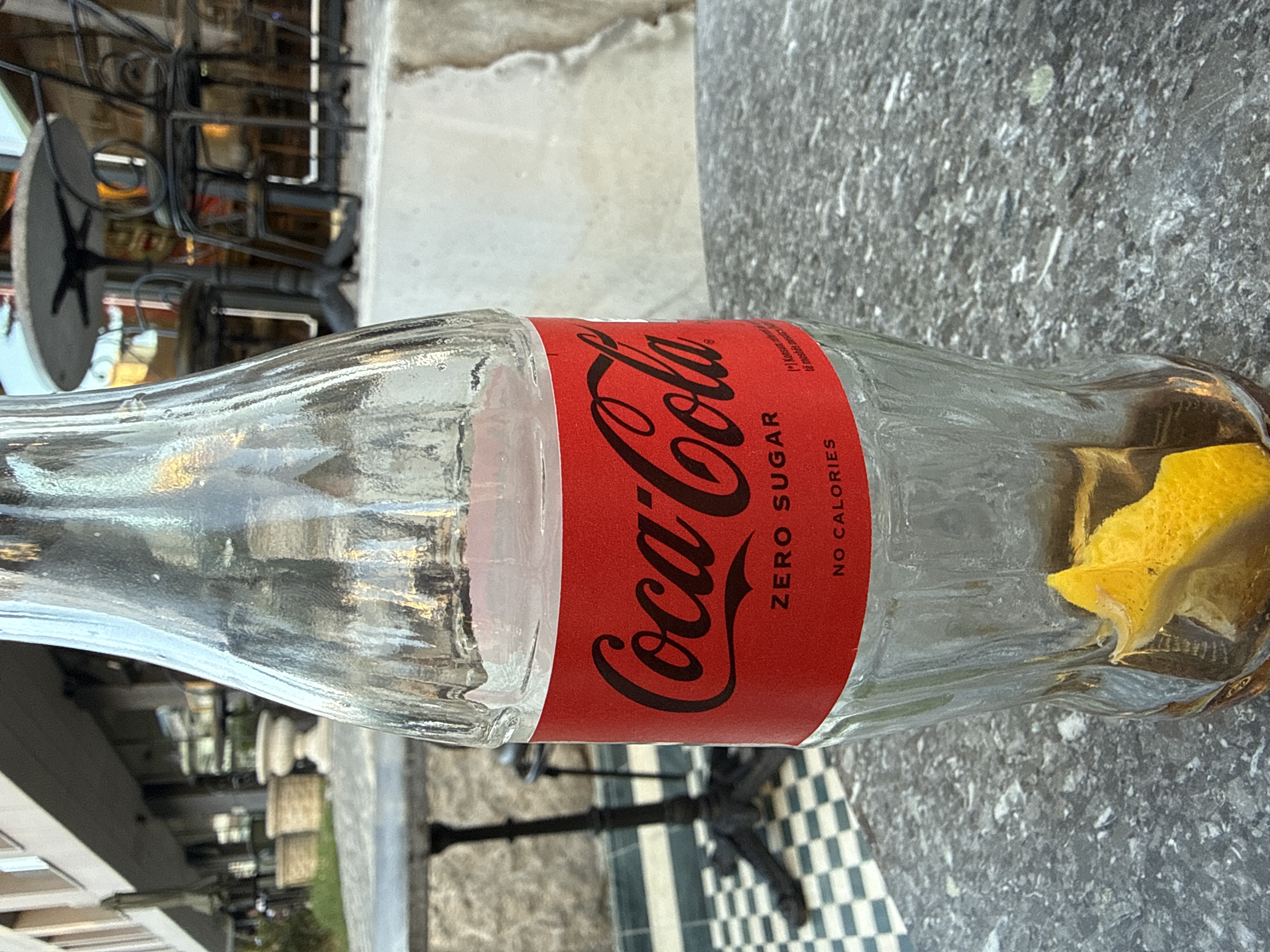 An empty Coca-Cola Zero Sugar glass bottle with a lemon wedge inside, standing on a marble cafe table
